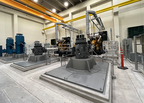 Interior view of a pump station with large vertical pumps, engines, pipes, metal platforms, railings, and an overhead crane inside an industrial facility.