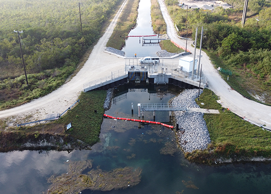 Aerial view of a pump station controlling water flow in a narrow canal, surrounded by gravel roads, riprap, and dense green vegetation.