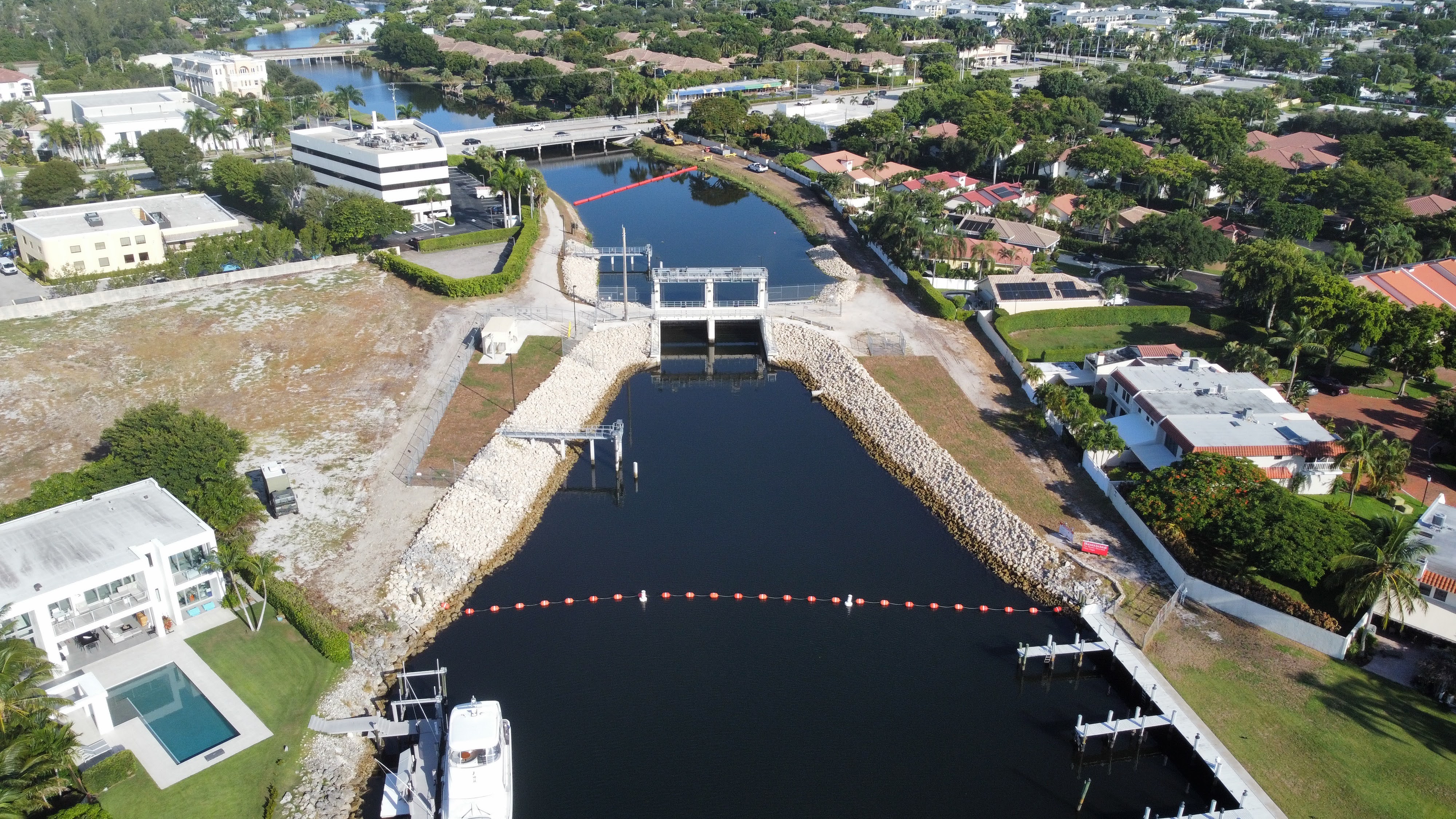 Aerial view of a canal flood control structure with riprap banks, floating barriers, docks, and nearby waterfront homes in an urban residential area.
